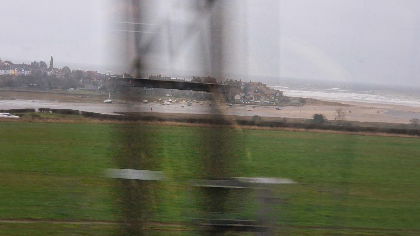 Across a field, by the beach, a charming village is visible on a small peninsula, surmounted by a cloudy grey sky.