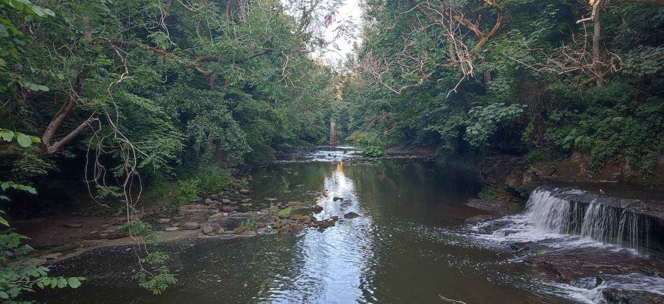 A view from the middle of a river — water pours down a dam on the right, while in the dead centre, a pillar is visible in the distance.