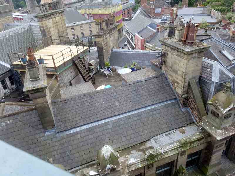 On the roof of a building in a crowded city, some chairs, a plastic table, and a ladder up to a small wooden platform.